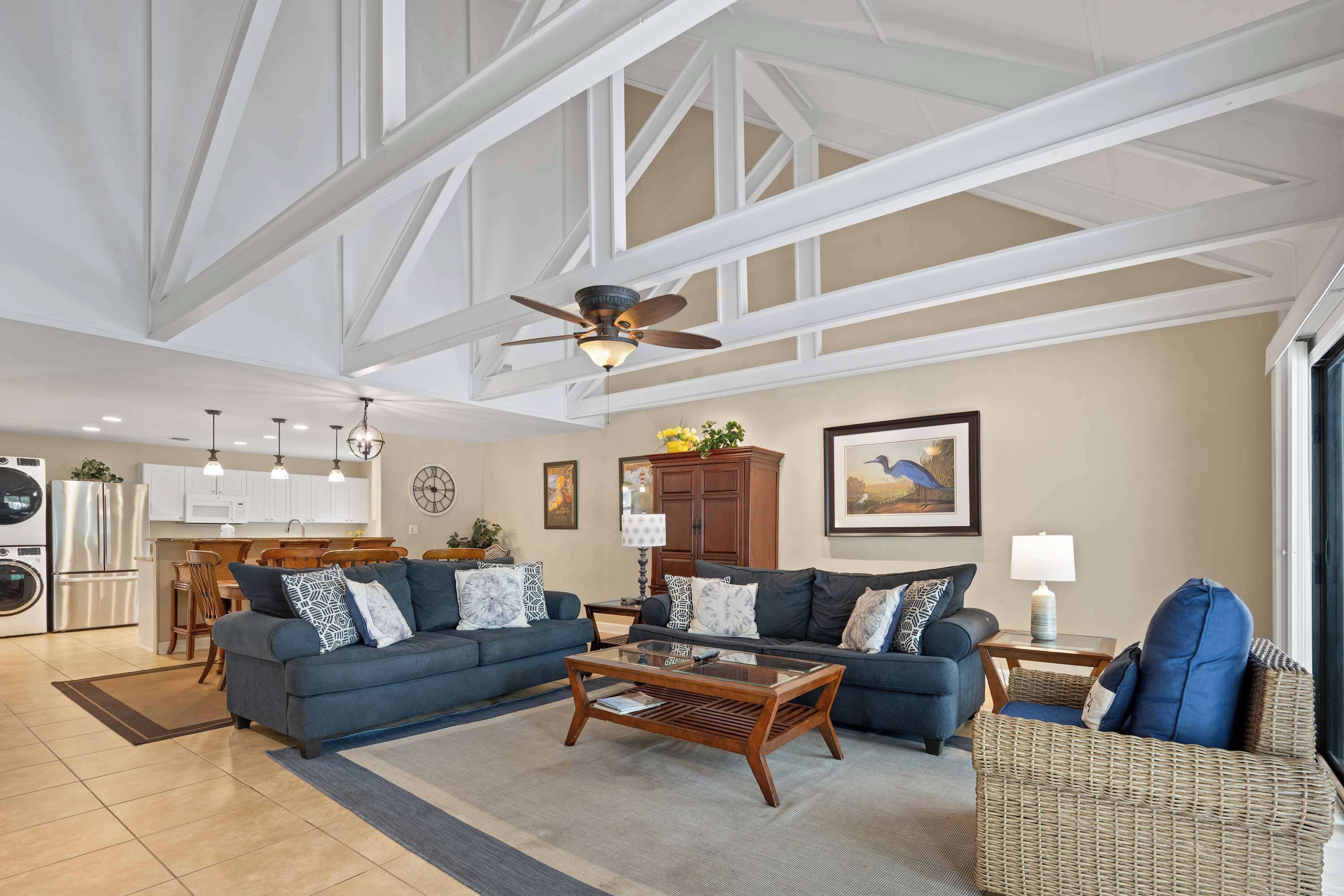A spacious living area with a high ceiling, a ceiling fan, a kitchen in the background, and a large window.