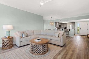 Ein Wohnzimmer mit einer Eckcouch, einem runden Couchtisch, einem Beistelltisch mit einer Lampe und einer Küche im Hintergrund.