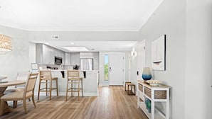 A modern kitchen with white cabinets, a dining area with wooden chairs, and a hallway with a wicker console. - Coastal condo on the beach with panoramic views, beach access & a shared pool (Hilton Head)