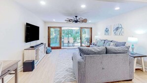 A modern living room with a grey sofa, a flat-screen TV, and a ceiling fan. - Sailmaster Commons, Unit 16  Sun soaked villa with pool access and beach nearby (Hilton Head)