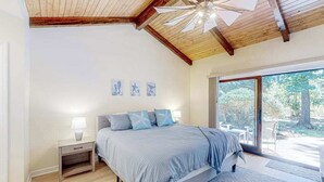 A bedroom with a large bed, a nightstand, a ceiling fan, and a view of the outdoors. - Sailmaster Commons, Unit 16  Sun soaked villa with pool access and beach nearby (Hilton Head)