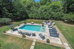 A rectangular pool with lounge chairs and umbrellas, surrounded by a grassy area and trees.
