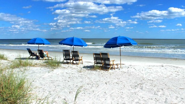 Ein Strand mit drei blauen Sonnenschirmen und Liegestühlen.