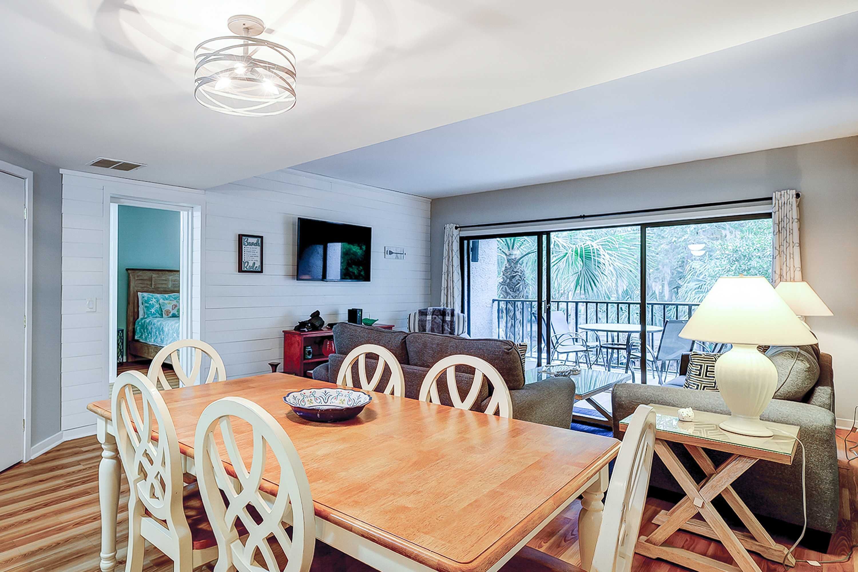 Un comedor con una mesa y sillas de madera, un televisor montado en la pared y un ventanal con vistas a palmeras.