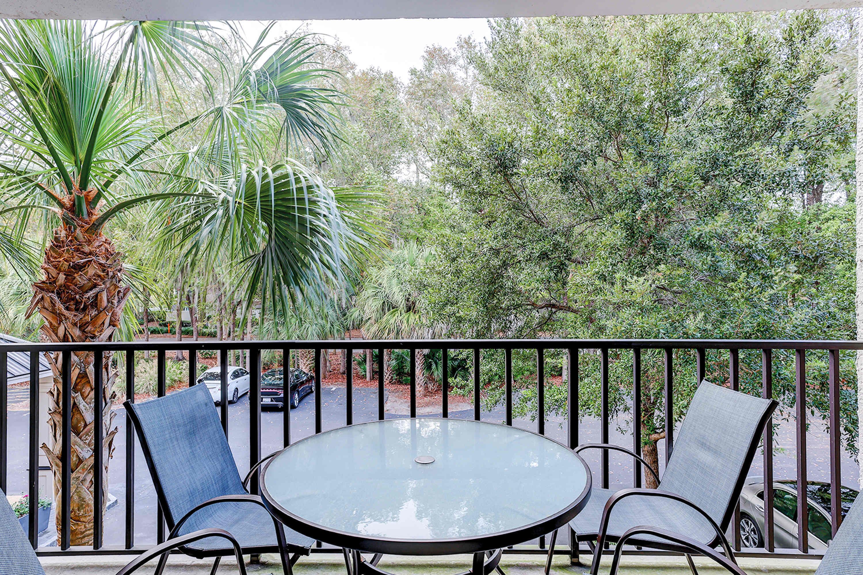 A balcony with a glass table and chairs, surrounded by a metal railing, overlooking a street with parked cars and trees.