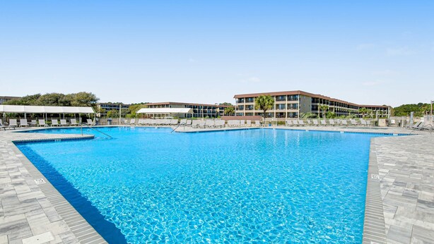Uma grande piscina ao ar livre com superfície de água azul e límpida, rodeada por um deck ladrilhado e um edifício com várias varandas.