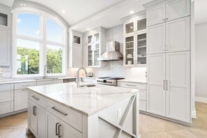 A modern kitchen with white cabinetry, a large island, a stove, and a window providing a view of greenery. - Stunning oceanfront home with pool, private boardwalk, decks, & elevator (Hilton Head)