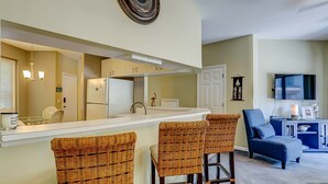 A kitchen with a white countertop, a refrigerator, and a sink. There are three wicker chairs and a blue armchair in the living area. - Dog-friendly island escape near the beach with pool, water view & WiFi (Hilton Head)