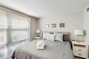 A neatly made bed with a folded towel, a bedside table with a lamp, and a window with blinds. - Lagoon View Villa 2 Pools walk to beach & shops (Hilton Head)