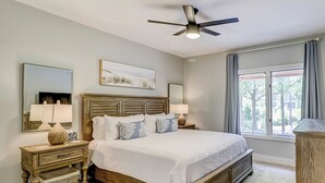 Une chambre avec un grand lit, deux tables de chevet, un ventilateur de plafond, une fenêtre avec des rideaux et un tableau au mur.
