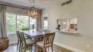 Un coin repas avec une table et des chaises en bois, un luminaire suspendu, une cuisine dotée d’un plan de travail et d’un évier, et une grande fenêtre avec des rideaux.