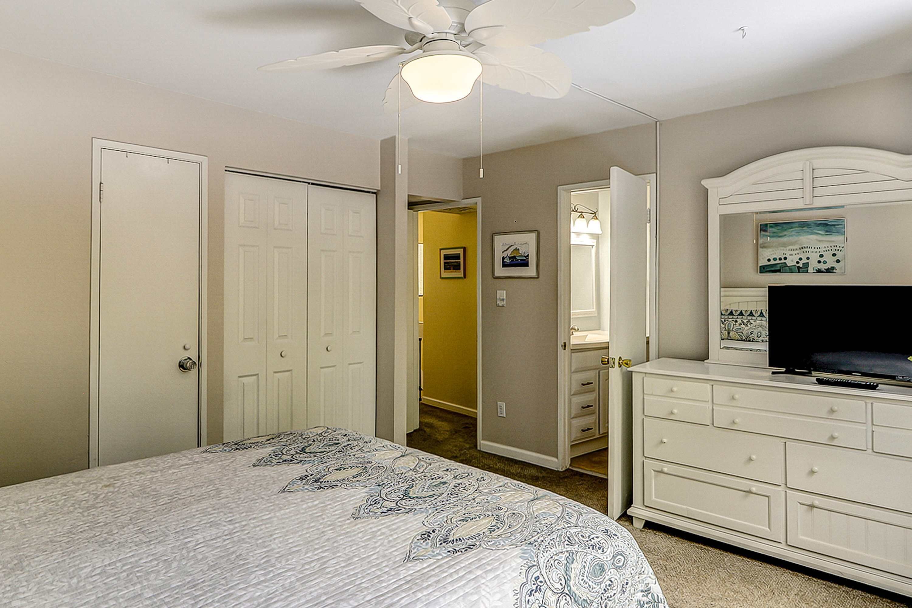 A bedroom with a bed, a dresser, a television, a ceiling fan, and a hallway leading to another room.