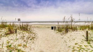 Ein Sandweg am Strand, der zum Meer führt, mit hohem Gras und bewölktem Himmel.