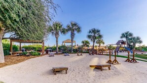 Zona de playa con arena, bancos de madera, un pabellón, palmeras y un área de juegos.