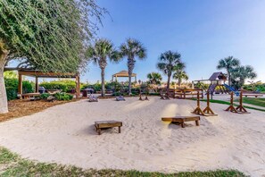 Ein Sandstrand mit Holzliegen, einer Laube, Palmen und einem Spielplatz.