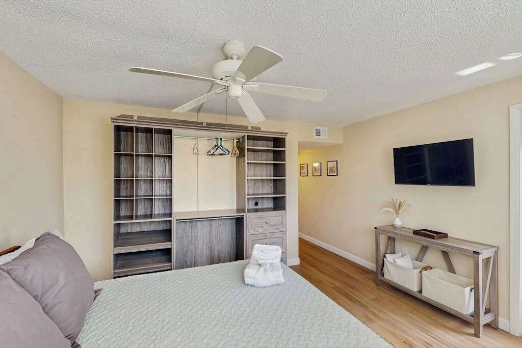 Quarto com cama, ventoinha no teto, televisão, guarda-roupa e mesa consola.