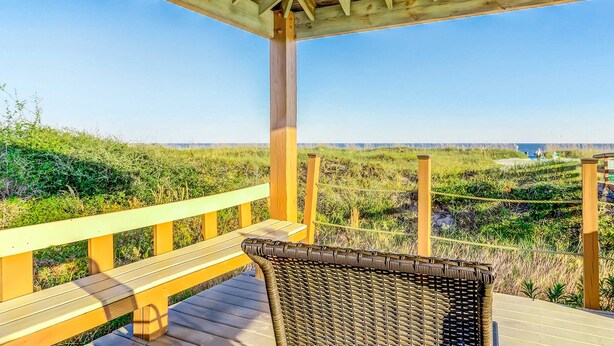 Una terraza de madera con una silla de mimbre, con vistas a una playa con dunas y cielo despejado.