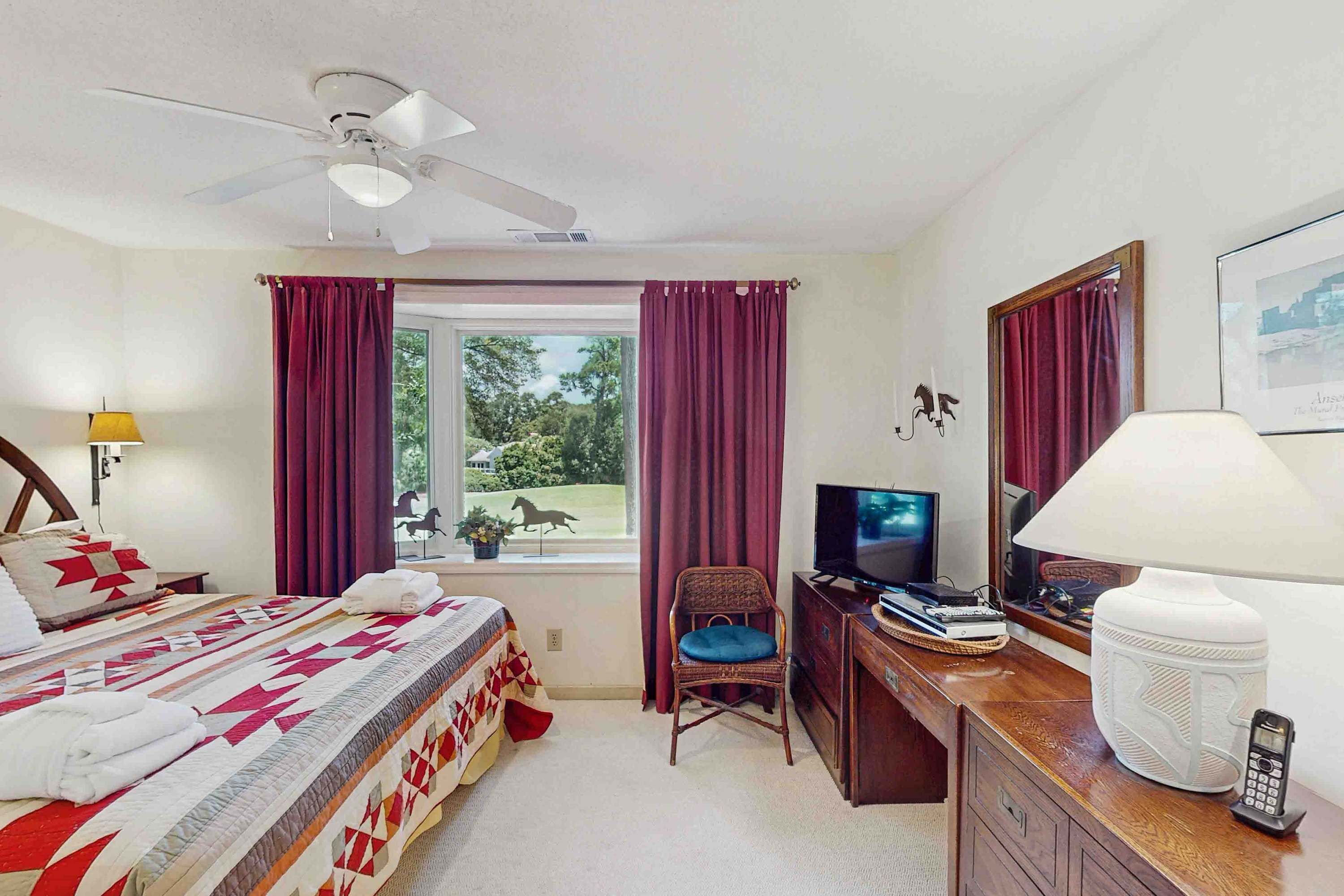 A hotel room with a bed, a desk with a TV, a chair, a ceiling fan, and a window with curtains.