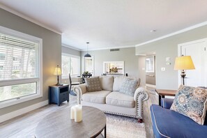 A living room with a sofa, a coffee table, and a lamp. There is a window with blinds, a door, and a kitchen visible in the background. - Total renovated Villa June 2024, views, pool, hot tub & beach club pick up (Hilton Head)