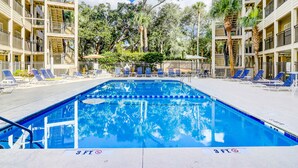 Una piscina con divisiones azules para carriles, rodeada de tumbonas y palmeras, con un edificio de varios pisos al fondo.