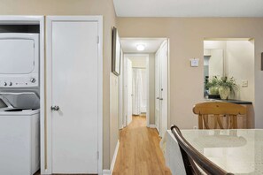 A hallway with a wooden floor, a white door, a white washing machine, a dining table with chairs, and a mirror.