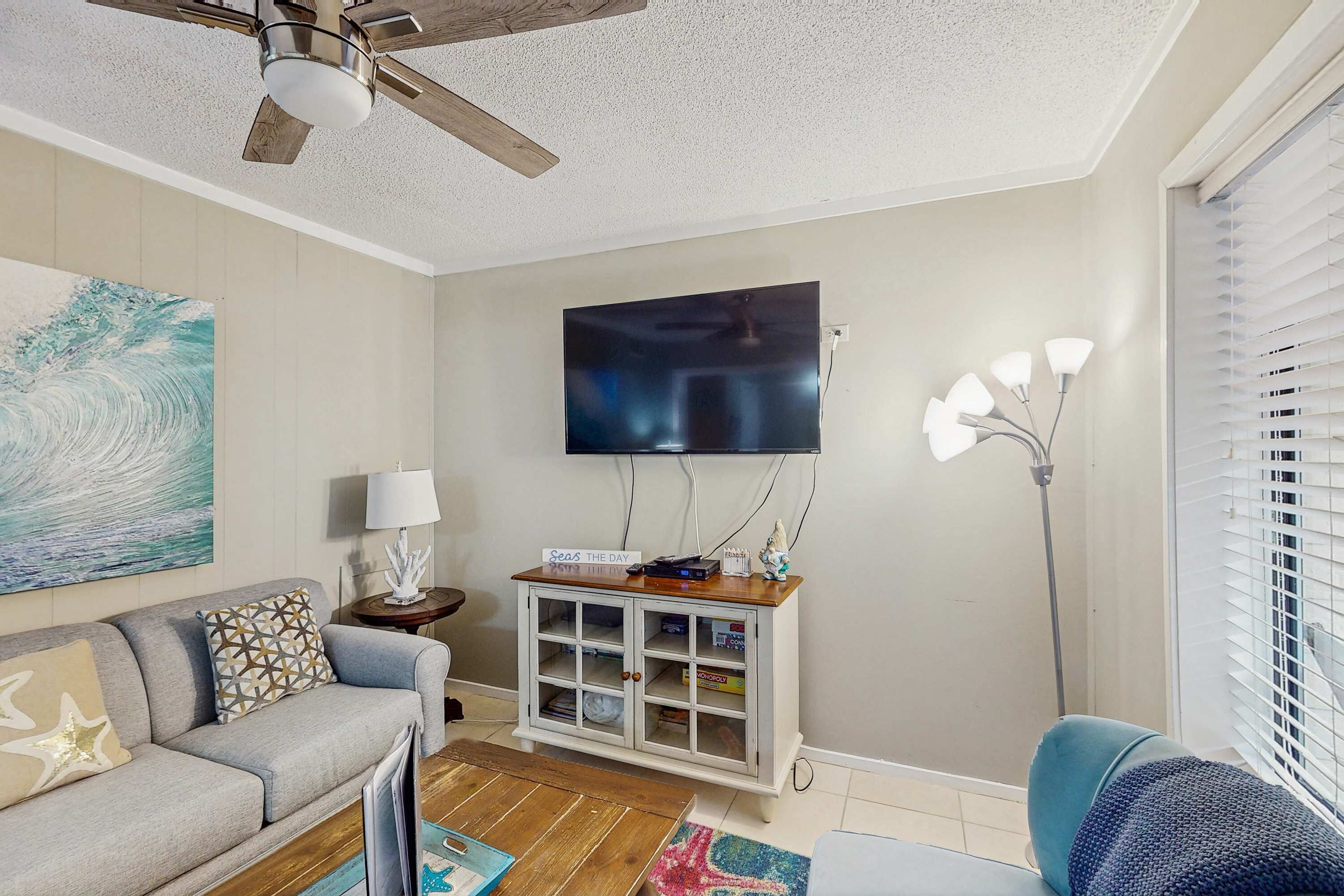 Een woonkamer met een plafondventilator, een flatscreen televisie aan de muur, een bank met decoratieve kussens, een bijzettafeltje met een lamp en een decoratiemöbel.