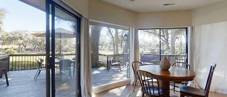 A sunlit room with a large sliding glass door leading to an outdoor deck, a round wooden table with chairs, and a rug on the floor.