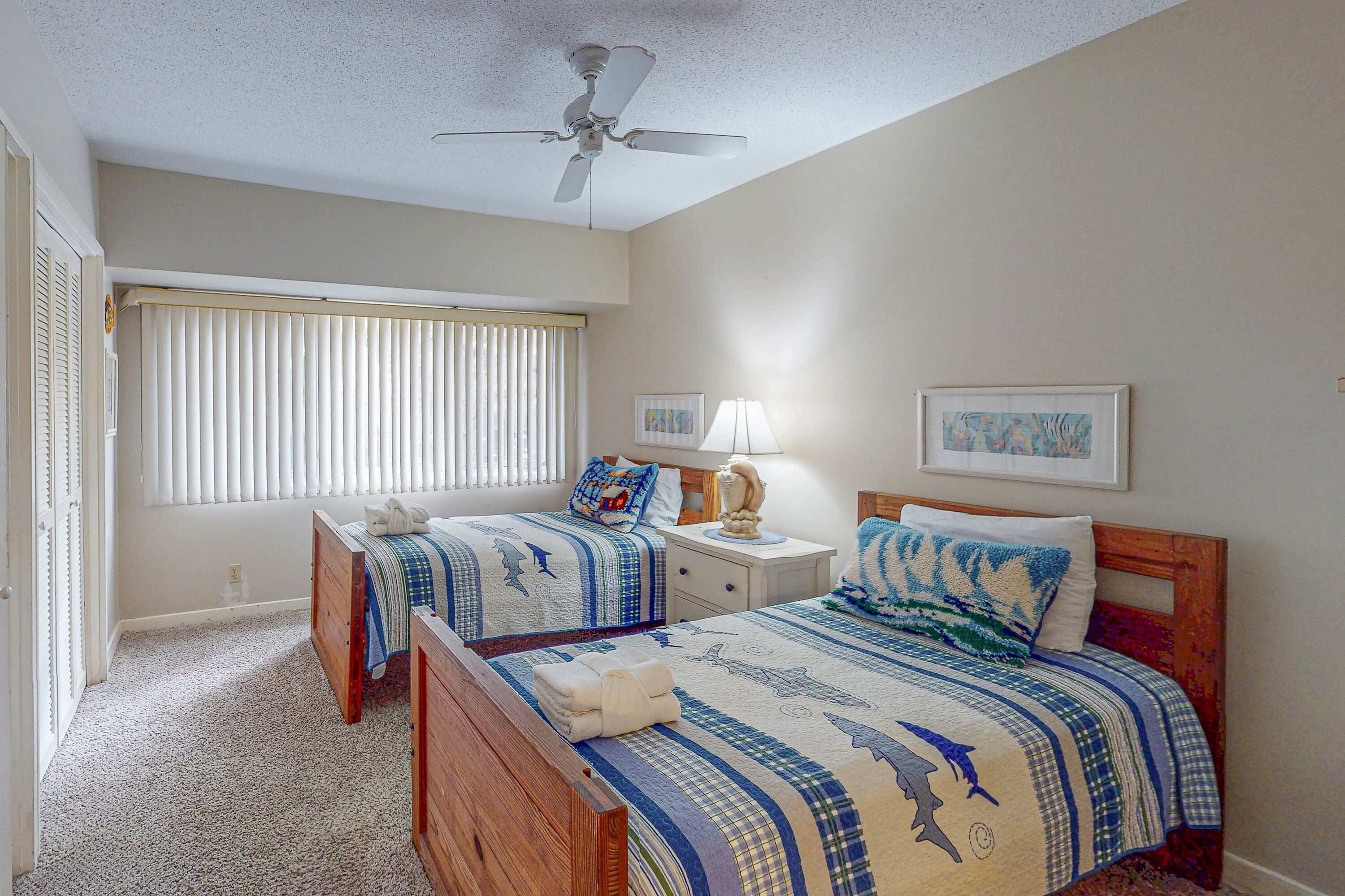 A bedroom with two beds, a ceiling fan, a window with blinds, a nightstand with a lamp, and two framed pictures on the wall.