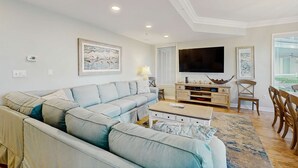 A living room with a sectional sofa, a coffee table, a TV, and a dining area with chairs. - Oceanfront condo with pools, hot tubs, WiFi, washer, dryer, & central AC (Hilton Head)