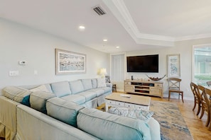 A living room with a sectional sofa, a coffee table, a TV, and a dining area with chairs. - Oceanfront condo with pools, hot tubs, WiFi, washer, dryer, & central AC (Hilton Head)
