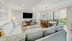 A modern living room with a sofa, coffee table, TV, and dining area. - Oceanfront condo with pools, hot tubs, WiFi, washer, dryer, & central AC (Hilton Head)