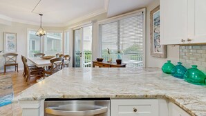 A kitchen with a marble countertop, white cabinets, a dining area with wooden chairs, and large windows with blinds. - Oceanfront condo with pools, hot tubs, WiFi, washer, dryer, & central AC (Hilton Head)