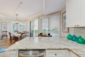 A kitchen with a marble countertop, white cabinets, a dining area with wooden chairs, and large windows with blinds. - Oceanfront condo with pools, hot tubs, WiFi, washer, dryer, & central AC (Hilton Head)