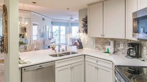 A kitchen with white cabinets, a dishwasher, a coffee maker, and a stove. - Beach dream with balcony view & shared pool, just steps away from the beach (Hilton Head)