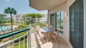 A balcony with a table and chairs, overlooking a swimming pool and a landscaped area with palm trees. - Beach dream with balcony view & shared pool, just steps away from the beach (Hilton Head)
