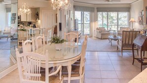 A spacious living area with a glass dining table surrounded by white chairs, a chandelier, and a view of the outdoors. - Beach dream with balcony view & shared pool, just steps away from the beach (Hilton Head)