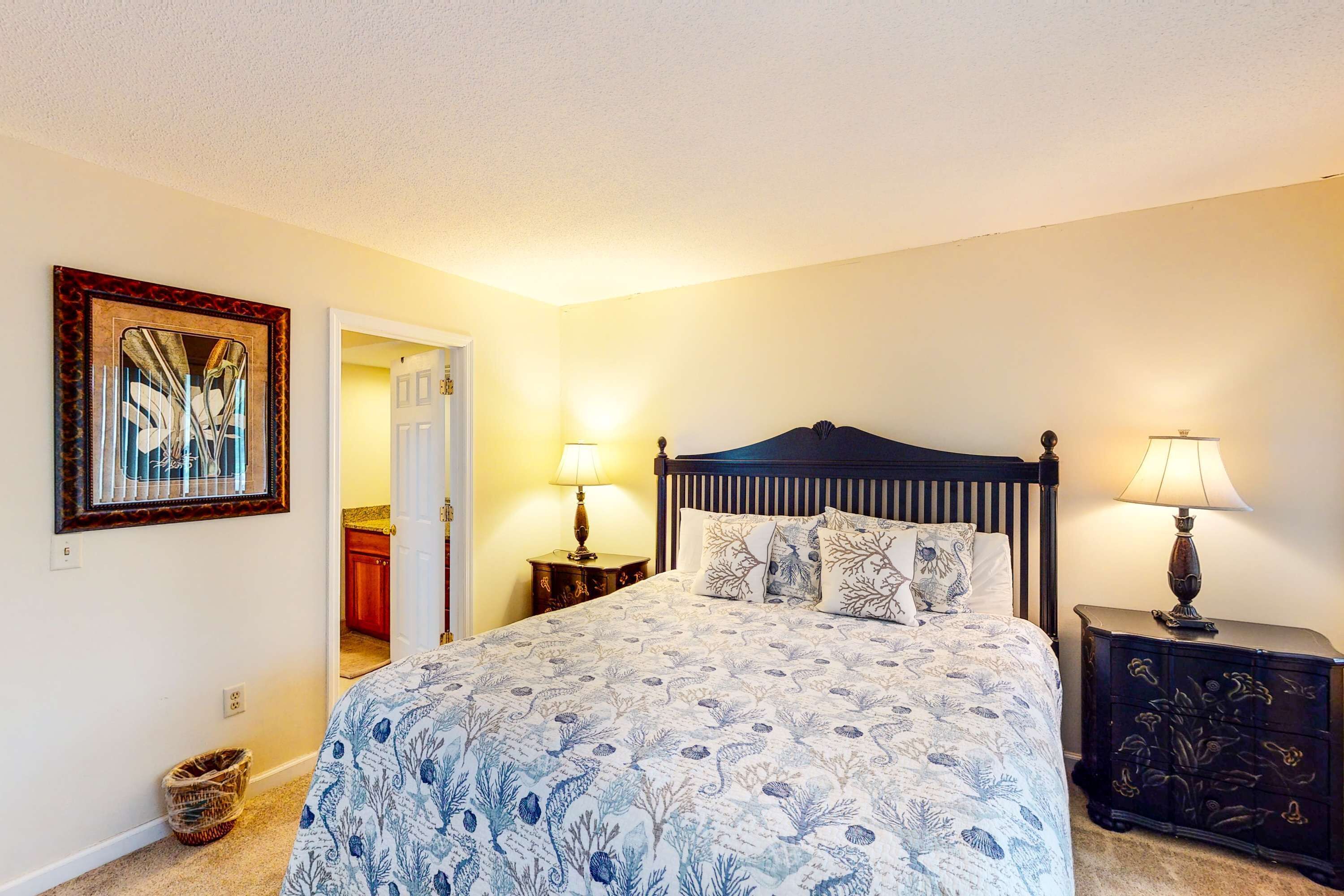 A bedroom with a bed, two bedside tables, a wall-mounted lamp, a door leading to another room, and a framed picture on the wall.
