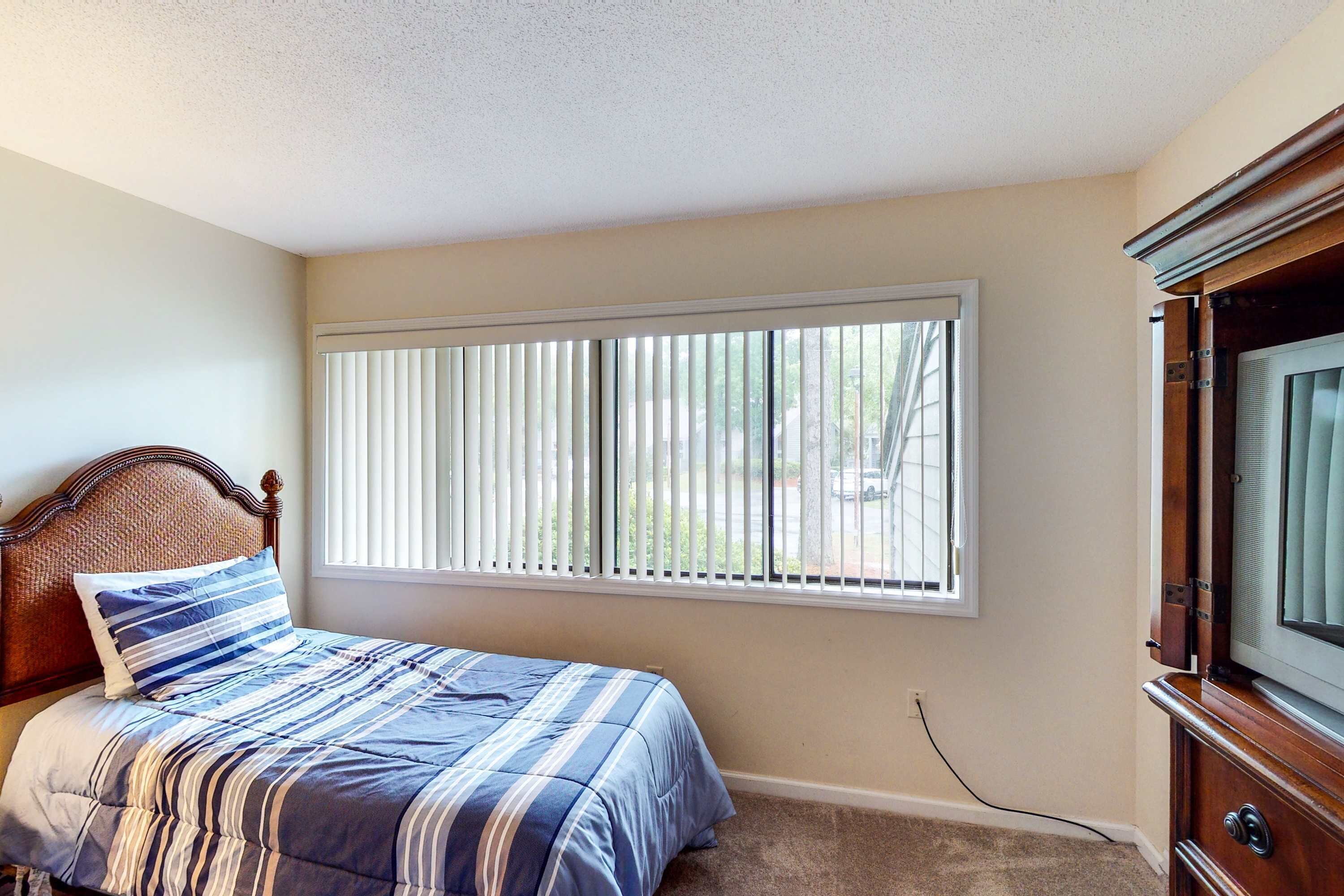 Une chambre à coucher avec un lit, une fenêtre avec des stores, un téléviseur et un sol recouvert de moquette.
