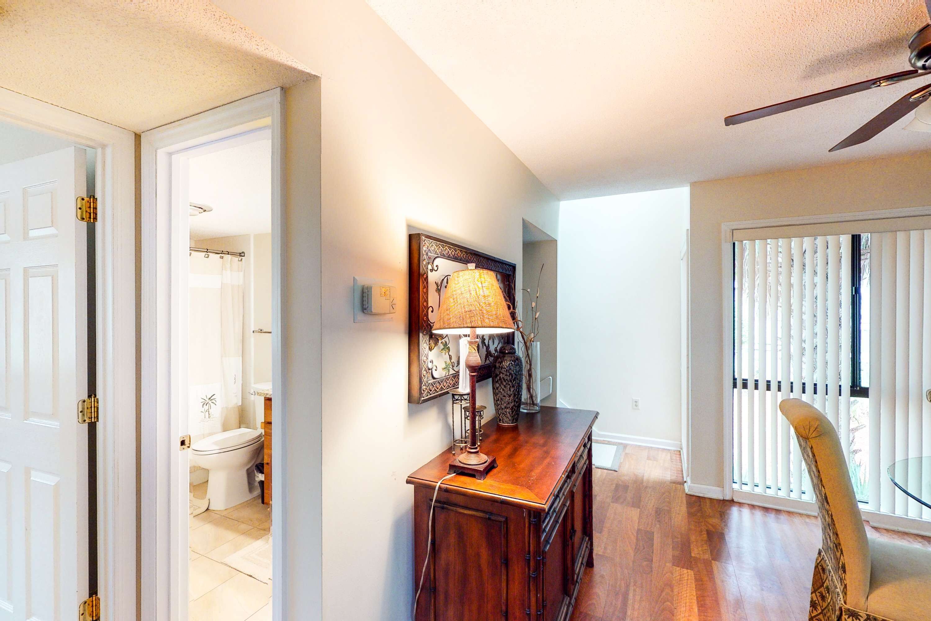 A hallway with a wooden desk, a lamp, and a chair. There is a door leading to a bathroom.