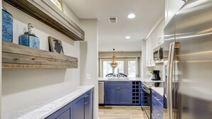 Een keuken met blauwe kastjes, een roestvrijstalen koelkast, een houten plankje met decoratieve objecten en een raam met natuurlijk licht.