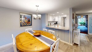 Un comedor con una mesa de madera, sillas, una araña y una cocina al fondo.