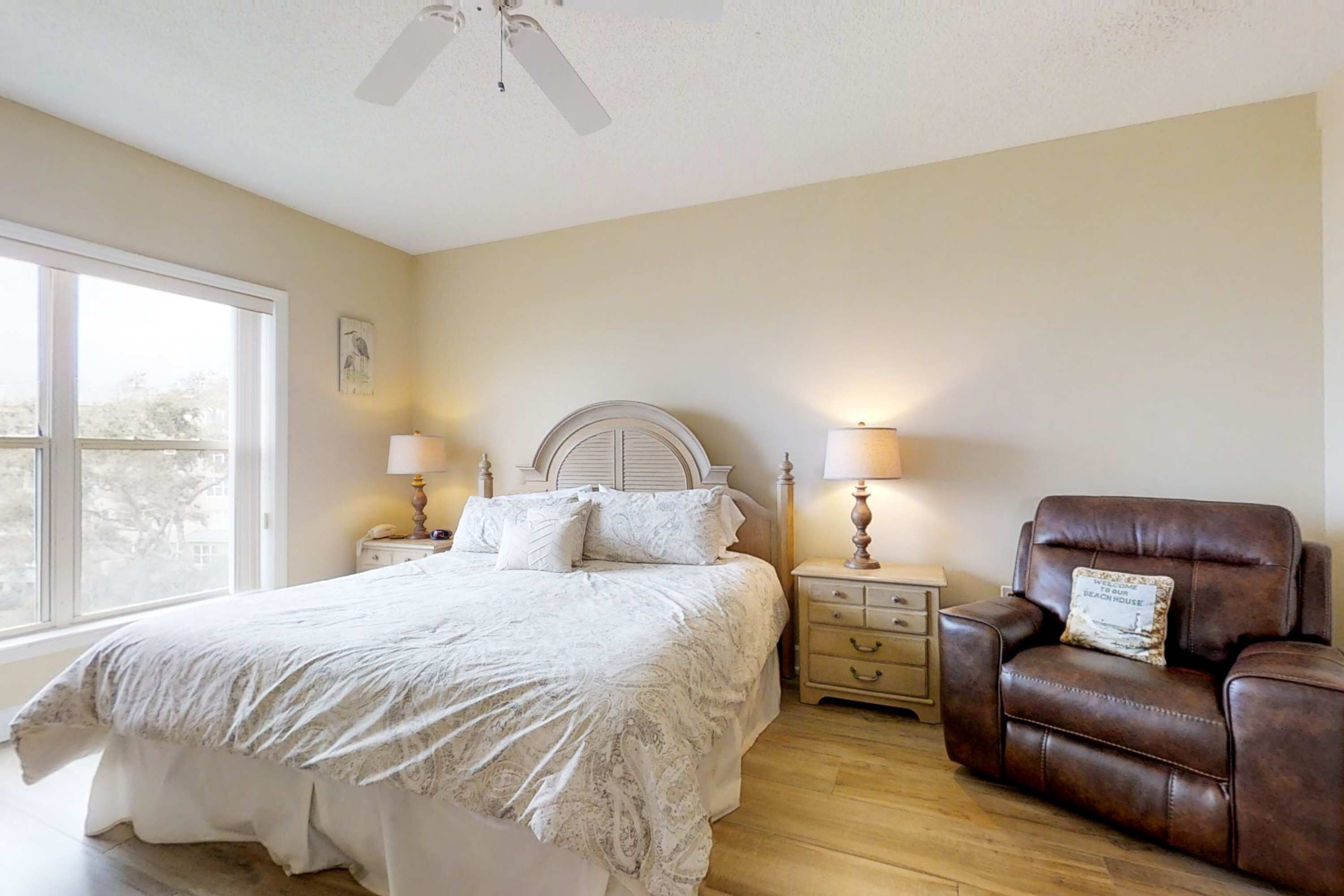 A bedroom with a bed, two bedside tables, a nightstand, a brown leather chair, and a window.