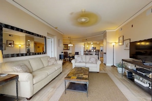 A spacious living room with two sofas, a coffee table, a TV, and a kitchen in the background. - Dreamy condo on the beach with balcony view, shared pool, & charming decor (Hilton Head)