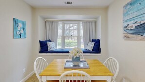 A dining area with a wooden table, white chairs, a window seat with blue cushions, and artwork on the walls. - Charming French Inspired Villa, Dog Friendly Retreat with Patio, Pool & Tennis (Hilton Head)