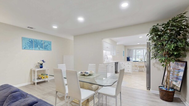 A modern living space with a glass dining table, white chairs, a sofa, a kitchen area, and a large potted plant. - Charming French Inspired Villa, Dog Friendly Retreat with Patio, Pool & Tennis (Hilton Head)