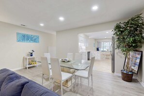 A modern living space with a glass dining table, white chairs, a sofa, a kitchen area, and a large potted plant. - Charming French Inspired Villa, Dog Friendly Retreat with Patio, Pool & Tennis (Hilton Head)