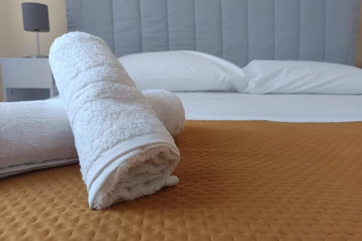 Un lit bien fait, avec une couette et des oreillers blancs, une serviette blanche roulée posée sur le lit et une lampe de chevet sur une table de nuit.