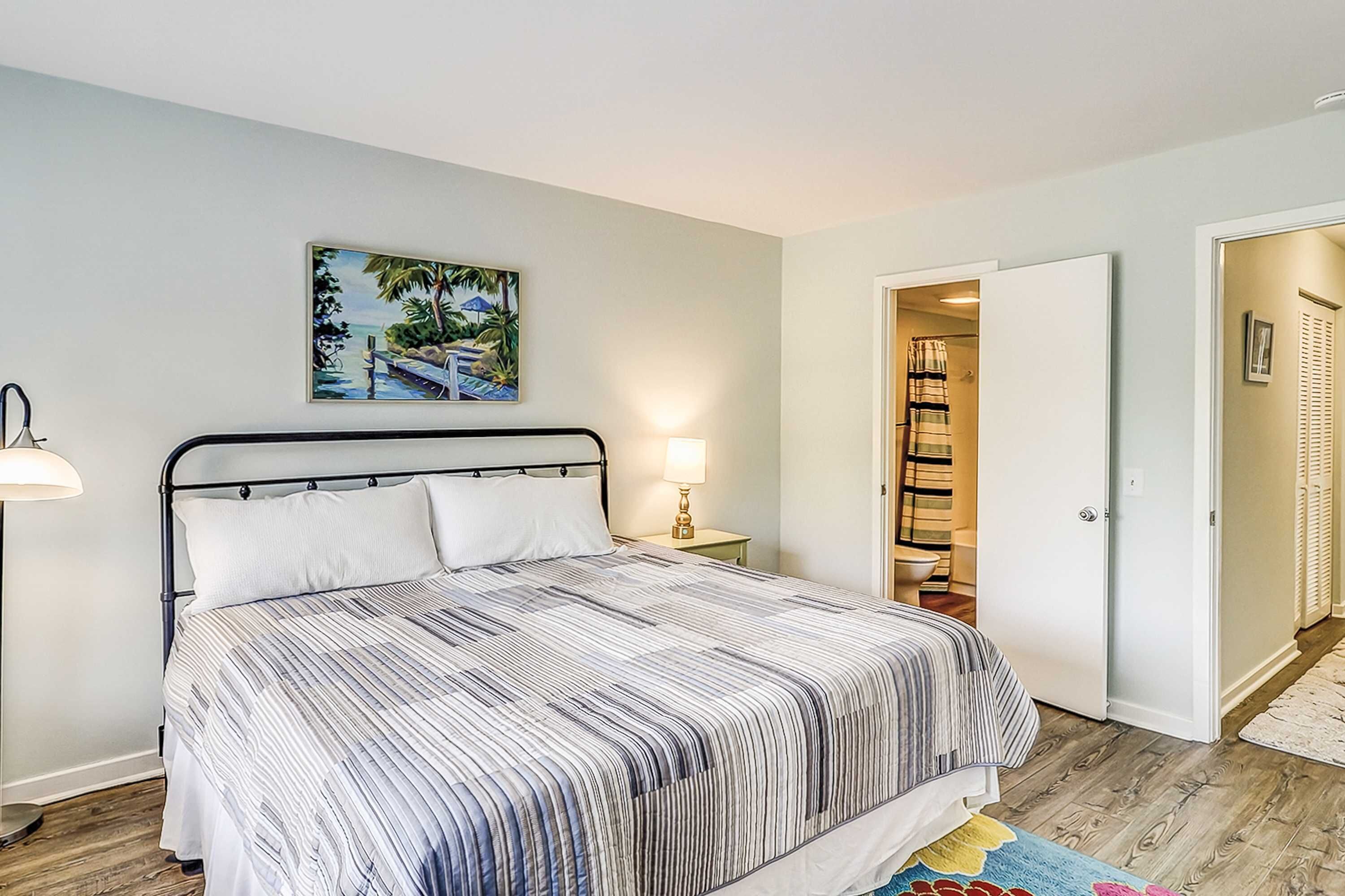 A bedroom with a bed, a nightstand, a lamp, a painting, a closet, and a door leading to another room.