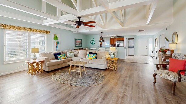 A spacious living room with a high ceiling, wooden beams, a ceiling fan, a large window with blinds, a sofa set, a coffee table, and a dining area with a table and chairs. - Gated community home with lagoon & pools Near Beaches & Dog Friendly (Hilton Head)