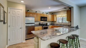 A kitchen with wooden cabinets, a stainless steel refrigerator, a microwave, a sink, and a granite countertop with bar stools. - Cozy dog-friendly home with free WiFi & private pool for a relaxing stay (Hilton Head)